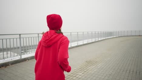 woman in red beanie and hoodie jogging along foggy pathway near iron railing, distant bridge with cars moving visible across railing, capturing active lifestyle and urban winter scenery