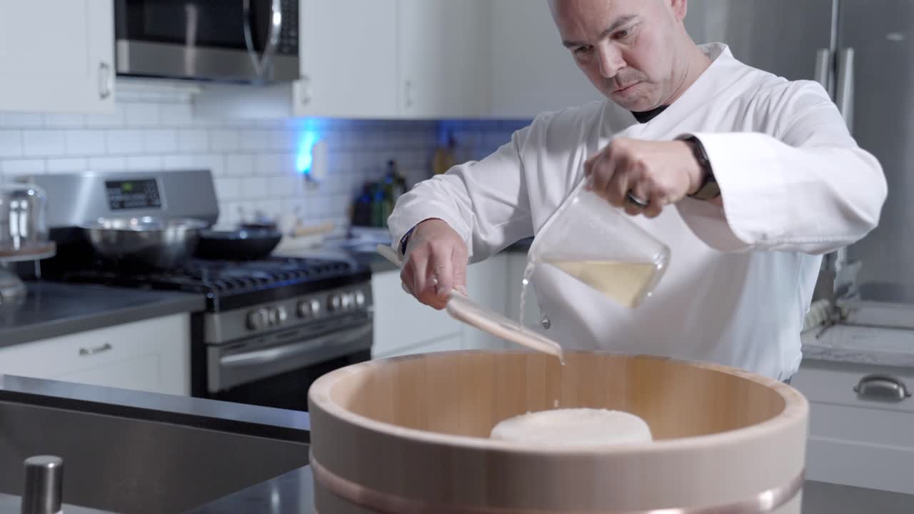 Wide-angle shot slides right as a sushi chef pours oil onto fresh rice in a wooden bowl, filmed at 120 fps to capture smooth motion and precision in culinary preparation.