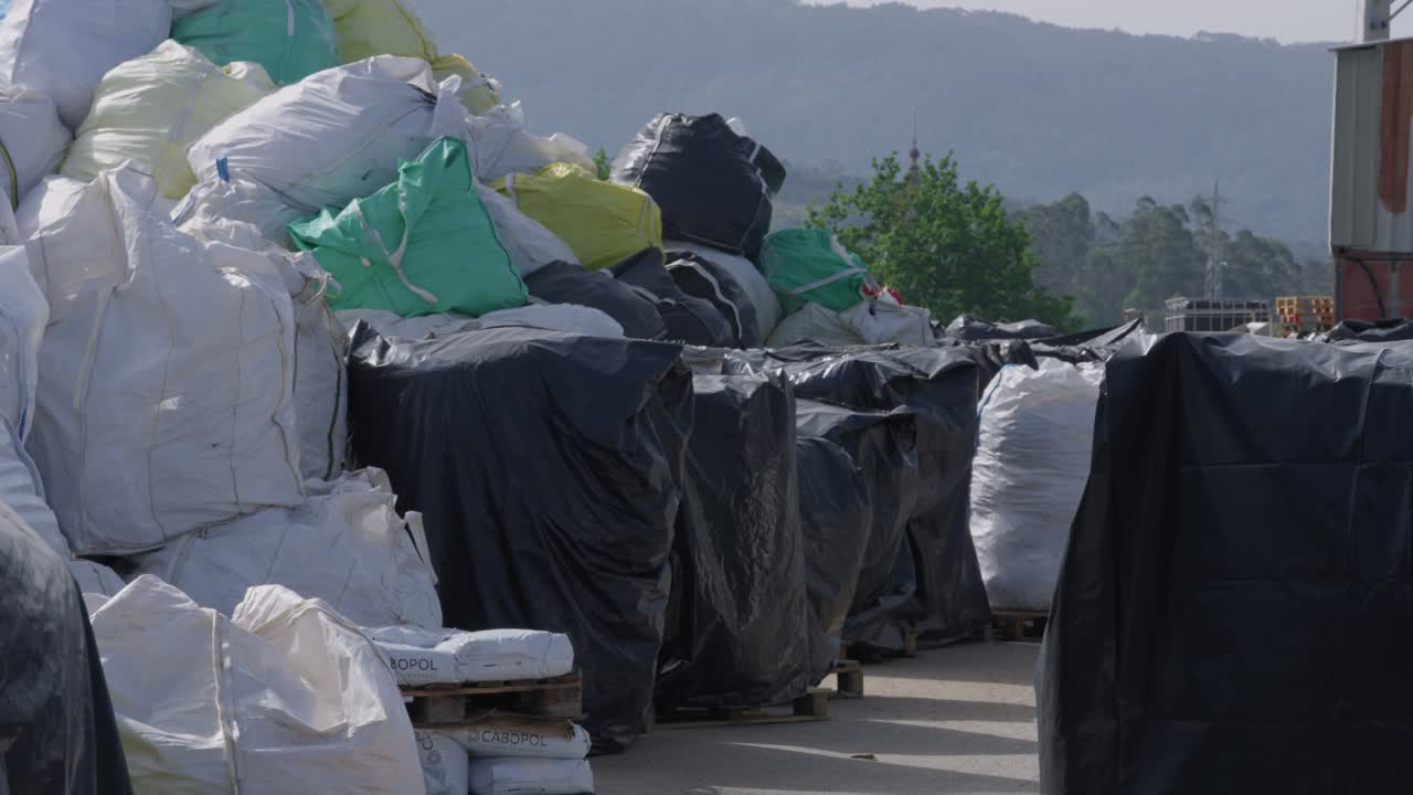 File di grandi sacchi di rifiuti di plastica e balle coperte su pallet impilati all'aperto in un impianto di riciclaggio con colline sullo sfondo