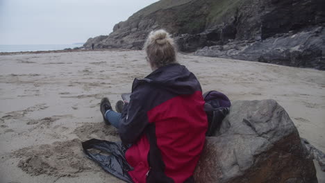 vista posterior de una mujer sentada en la arena con su tableta dibujando la vista costera en rinky head and cove en cornualles, inglaterra, reino unido