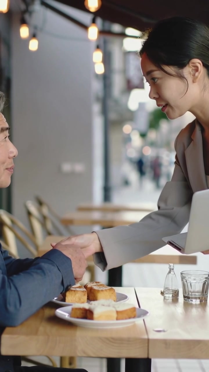Video verticale: Una donna in blazer arriva e gli stringe la mano al caffè, parlando di lavoro davanti a un laptop e dolci.