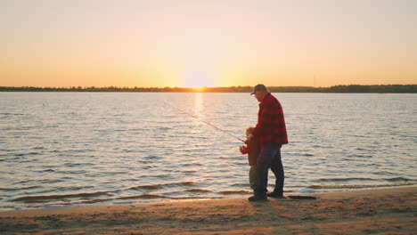 old fisher and his son or grandchild are catching fish by fishing rod on shore of lake or river