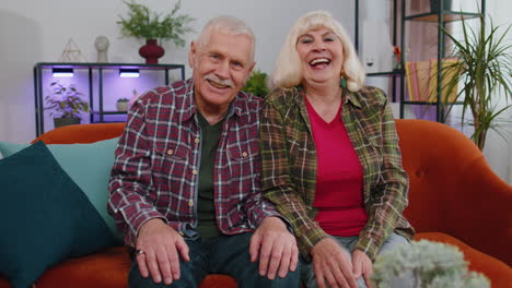 Happy-old-senior-elderly-family-couple-hugging,-laughing,-smiling-looking-at-camera-at-home-sofa