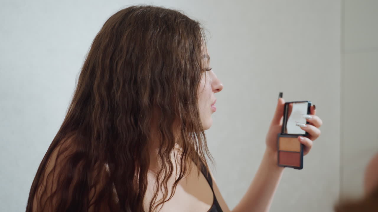 Mujer relajada con herramientas de maquillaje, mujer fotografiada aplicándose colorete y polvos con una brocha grande, mujer serena maquillándose con cabello texturizado y un ambiente de glamour delicado