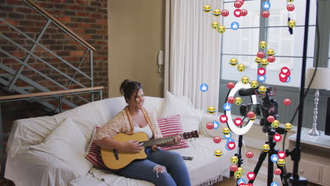 animation of emoji, love and like icons over female vlogger with guitar recording vlog at home