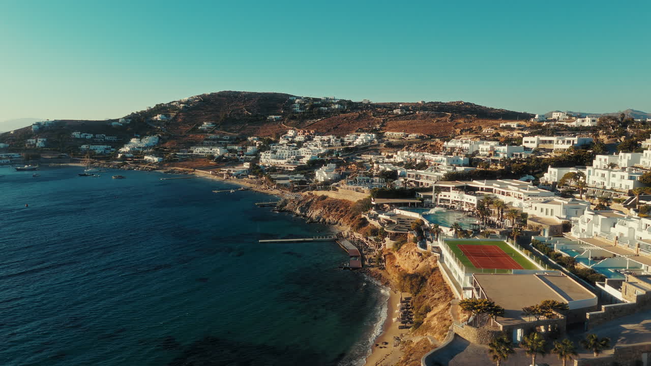 Drone orbiting and sliding sideways along the Mykonos coastline, showing white houses, rocky shore, and the blue Aegean Sea