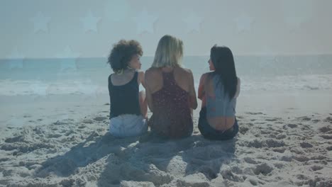 Multiple-star-icons-over-rear-view-of-three-diverse-female-friends-sitting-and-talking-on-the-beach