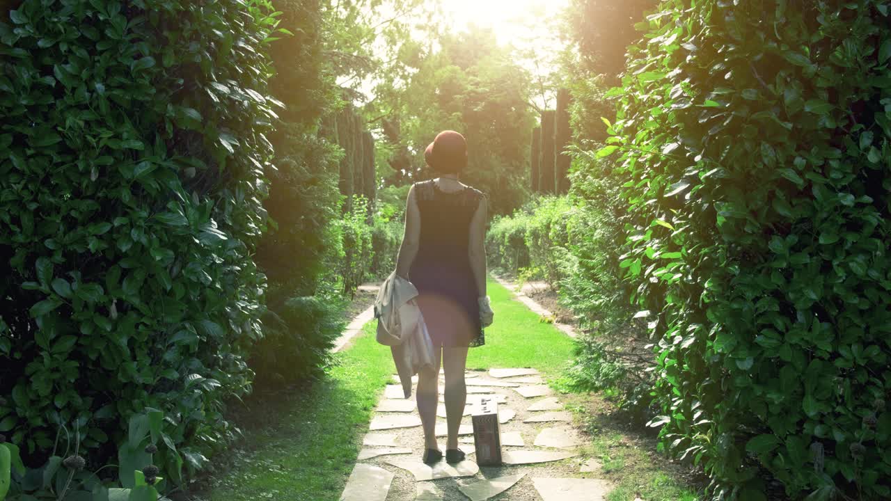 A person walks along a sun-drenched path in a lush green garden, carrying a suitcase.