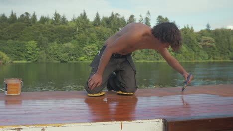 young man painting wooden boat cabin roof planking with protective exterior paint