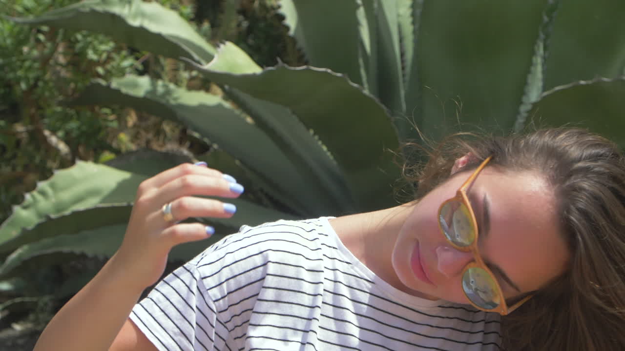 선글라스를 입은 젊은 여성이 아가베 식물 근처에 서서 아래를 바라보며 미소 짓고 있다