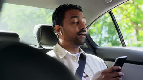 passenger with earphones and cellphone in taxi car.transport, business and technology concept  happy smiling indian male passenger with wireless earphones using smartphone on back seat of taxi car