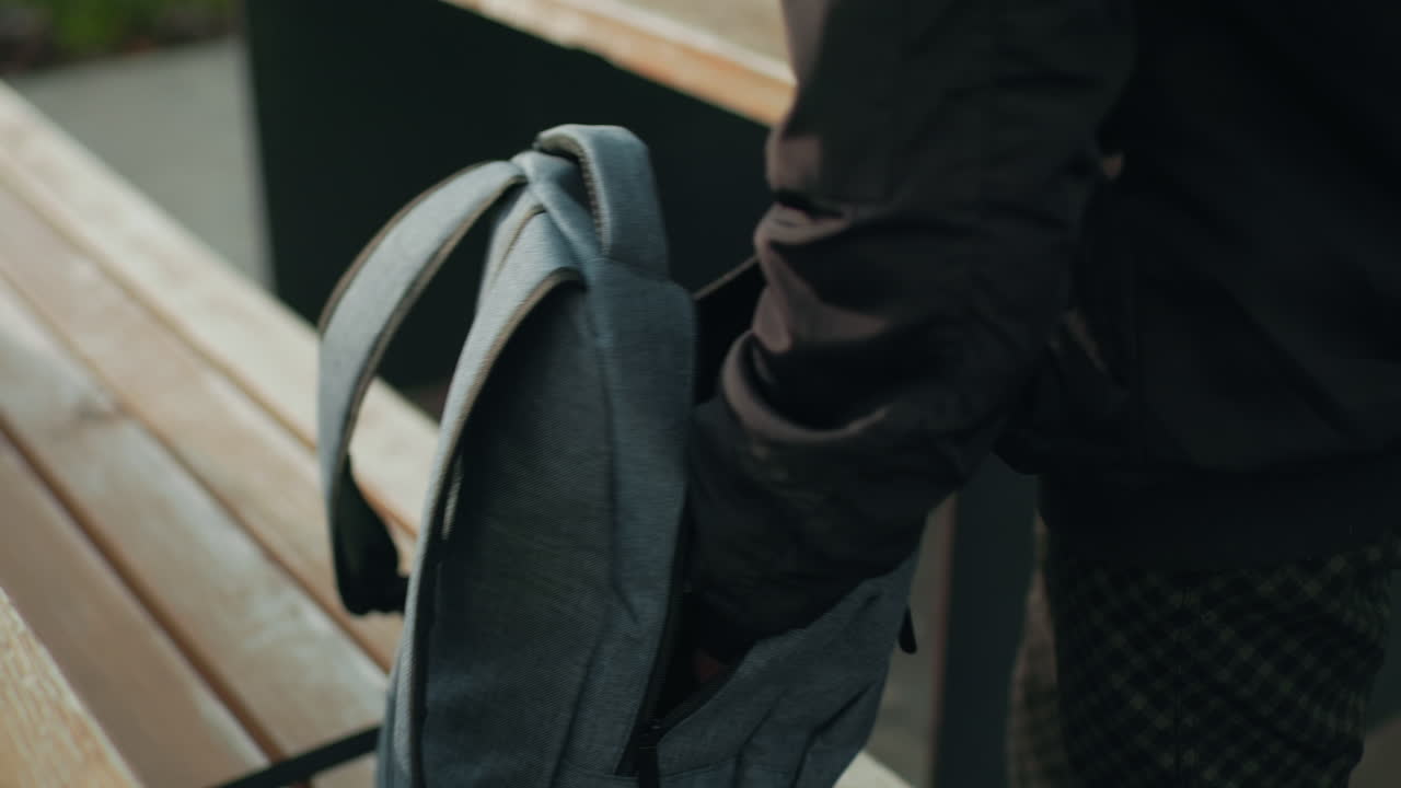 Close up of student removing book from backpack while standing beside wooden bench on paved outdoor area with natural daylight highlighting hands and fabric texture of bag during study preparation
