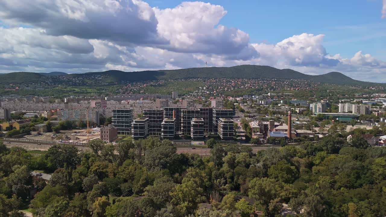 Vista orbital de drone de Óbuda, Budapeste, a partir da Ilha de Hajógyári, capturando o desenvolvimento urbano, prédios de apartamentos modernos e vegetação ao longo do Danúbio, com as colinas de Buda sob um céu azul nublado