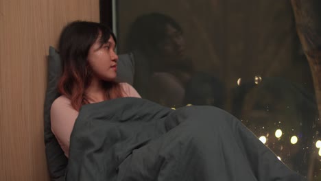 Sleepless Asian Girl Lying On Her Back Under Blanket and Looking Out The Window At Night