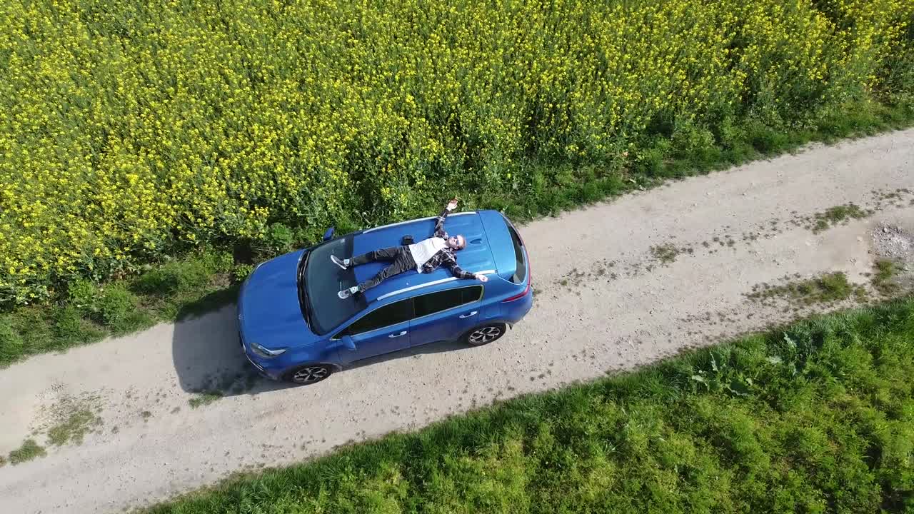 libertà interna di un uomo che si raffredda seduto sul tetto di una macchina blu in un campo giallo-verde di fiori - zoom out drone shot