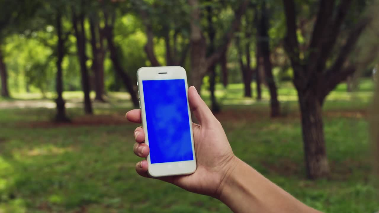 vista trasera de una mujer rubia sosteniendo un teléfono inteligente con pantalla azul y tecla de croma, navegando por el contenido. primer plano de un teléfono en manos de una chica en un fondo de parque. cámara lenta.