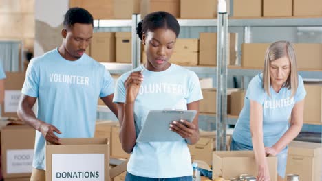 young african american female volunteer writing and checking donations in charity warehouse while her male coworkers sorting donated stuff in boxes in the background