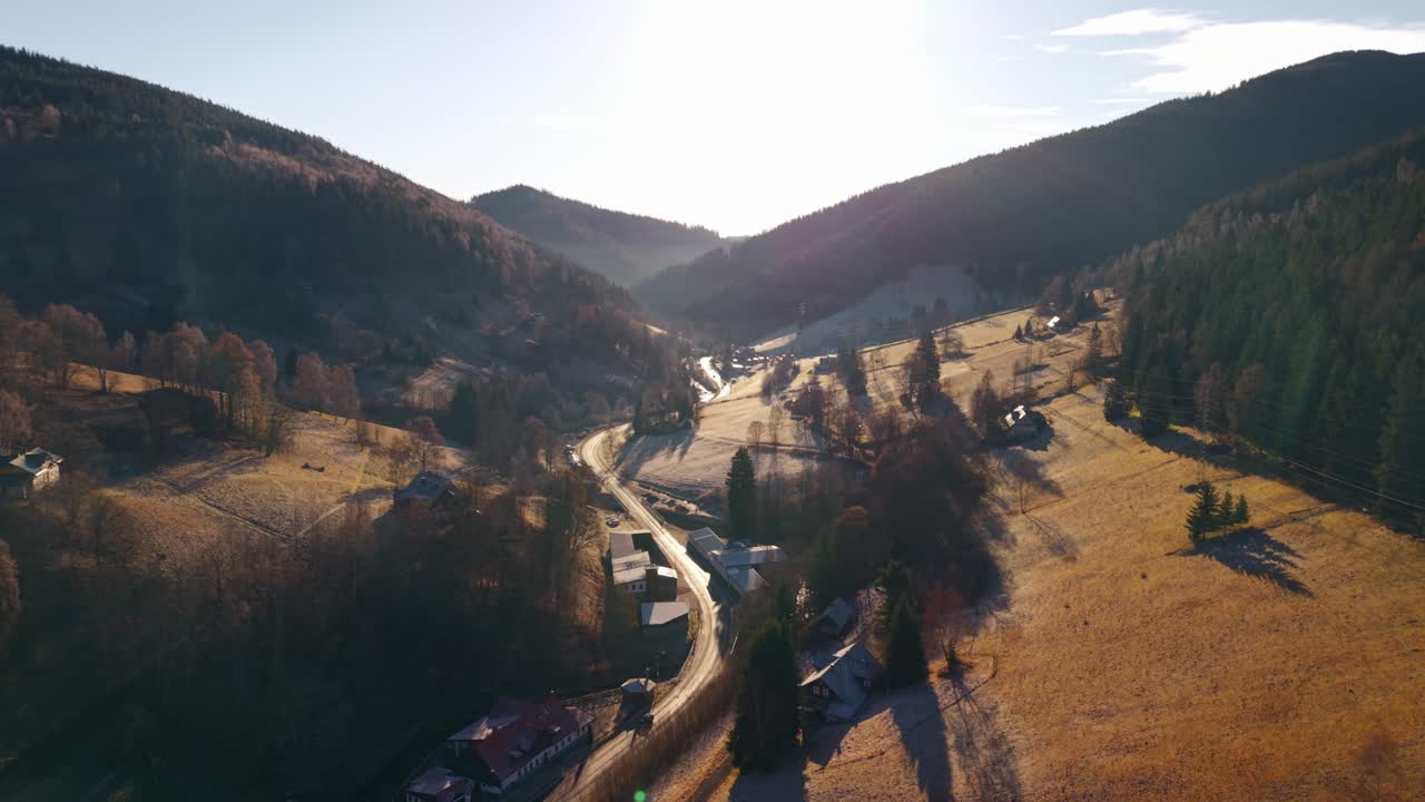 Vista aerea all'alba di una vallata montana con prati gelidi, colline boscose e villaggio rurale immerso nella calda luce del mattino