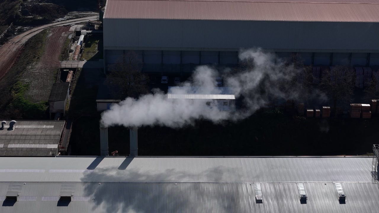 Active smokestacks emitting plumes of white smoke against a clear sky in sprawling factory complex nestled in a hilly landscape, with white smoke billowing from industrial chimney