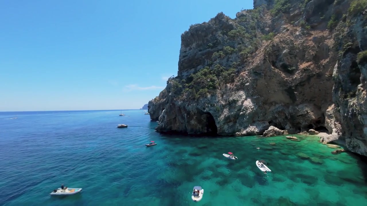 Små både flyder under tårnhøje kalkstensklipper og en havgrotte langs den barske kystlinje ved Cala Goloritze, Baunei, Sardinien — filmisk FPV droneoptagelse