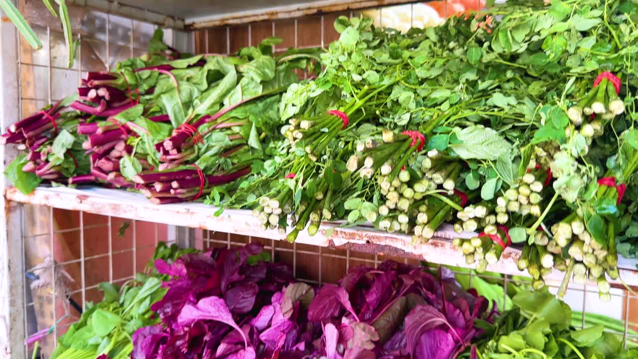 Camera pans down a market shelf displaying vibrant leafy greens, purple amaranth, and fresh herbs under bright natural lighting in an open-air setting