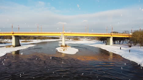 Yellow-bridge-over-river-Neris-in-Kaunas-city-during-snowfall,-aerial-low-angle-fly-backward-view