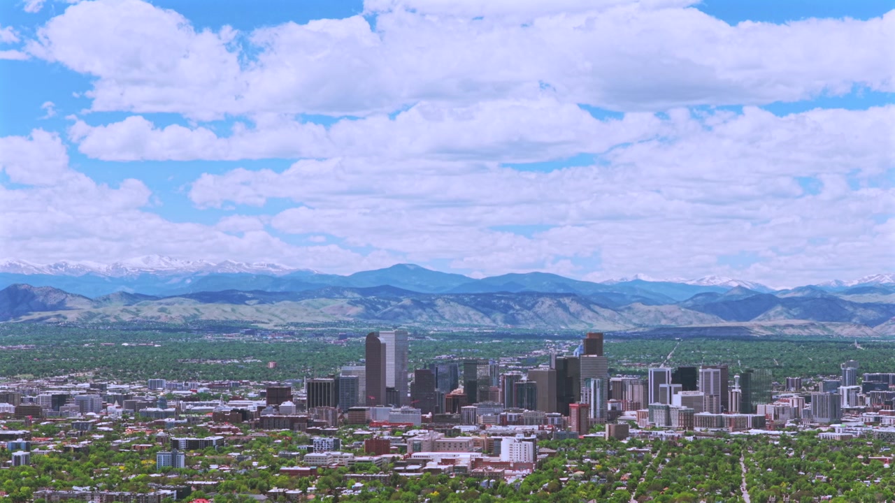 Denver, Colorado: Bybillede med skyskrabere og skyline i downtown, omgivet af Front Range's sneklædte Rocky Mountains. Droneoptagelse fra luften af forårskvarterer under en morgenblå himmel med skyer, der cirkler mod venstre.