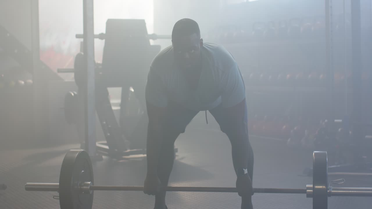 En afroamerikansk voksen mand ser en vægtstang med vægtskiver, griber stangen, bøjer sig forover og løfter for at styrketræne i fitnesscenteret.