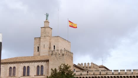 Toma-De-Abajo-Hacia-Arriba-Que-Muestra-La-Bandera-Española-Ondeando-En-El-Viento-En-La-Cima-De-La-Catedral-De-Mallorca-Contra-El-Cielo-Nublado