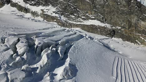 Vista-Aérea-De-Una-Llanura-De-Nieve-En-Los-Alpes-Suizos,-Cresta-De-Montaña-Rocosa-En-El-Fondo