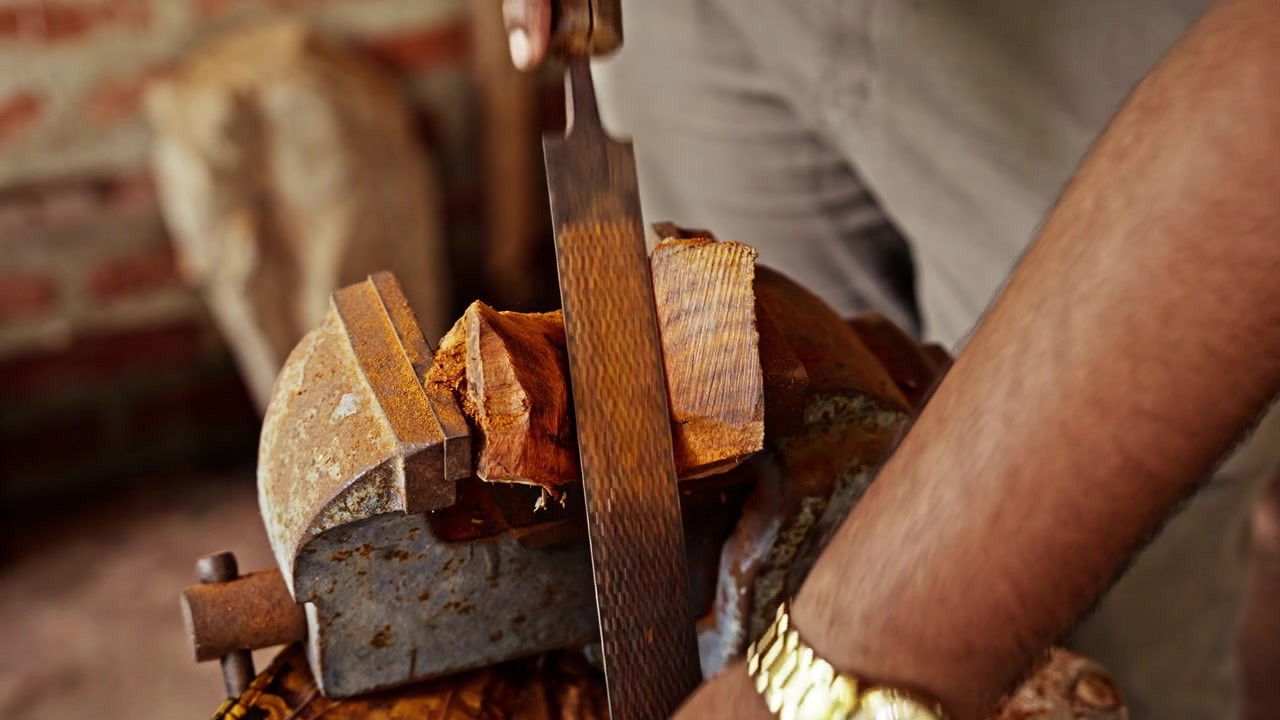 Um entalhador de madeira trabalha uma peça de madeira com uma lima manual em uma oficina de aldeia no Sri Lanka, onde entalhes e ferramentas preenchem a bancada. O ofício é passado de geração em geração.