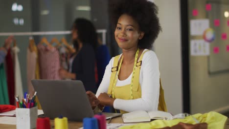 feliz diseñador afroamericano sentado frente a la computadora sonriendo con colegas en el fondo