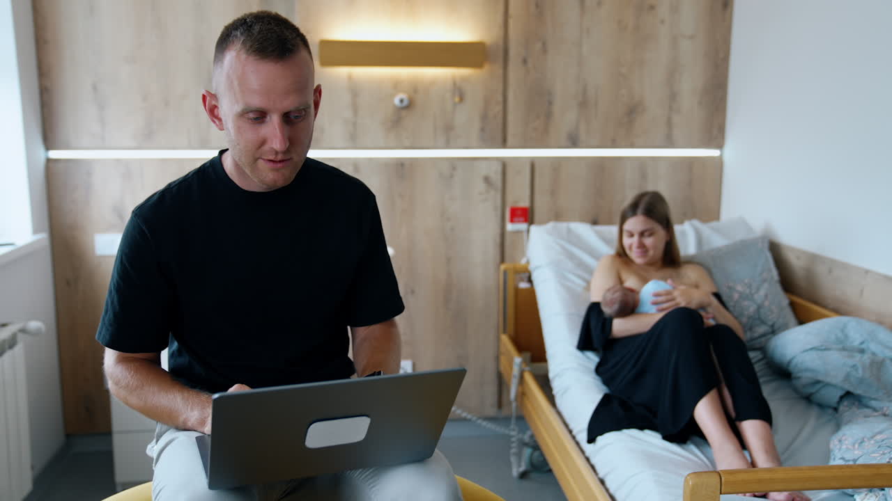 A new family of three. Parents with a newborn in a hospital. Father works at laptop while mother breastfeeding a child.