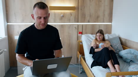 a new family of three. parents with a newborn in a hospital. father works at laptop while mother breastfeeding a child.