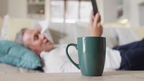 hombre caucásico feliz acostado en el sofá de la sala de estar, usando un teléfono inteligente