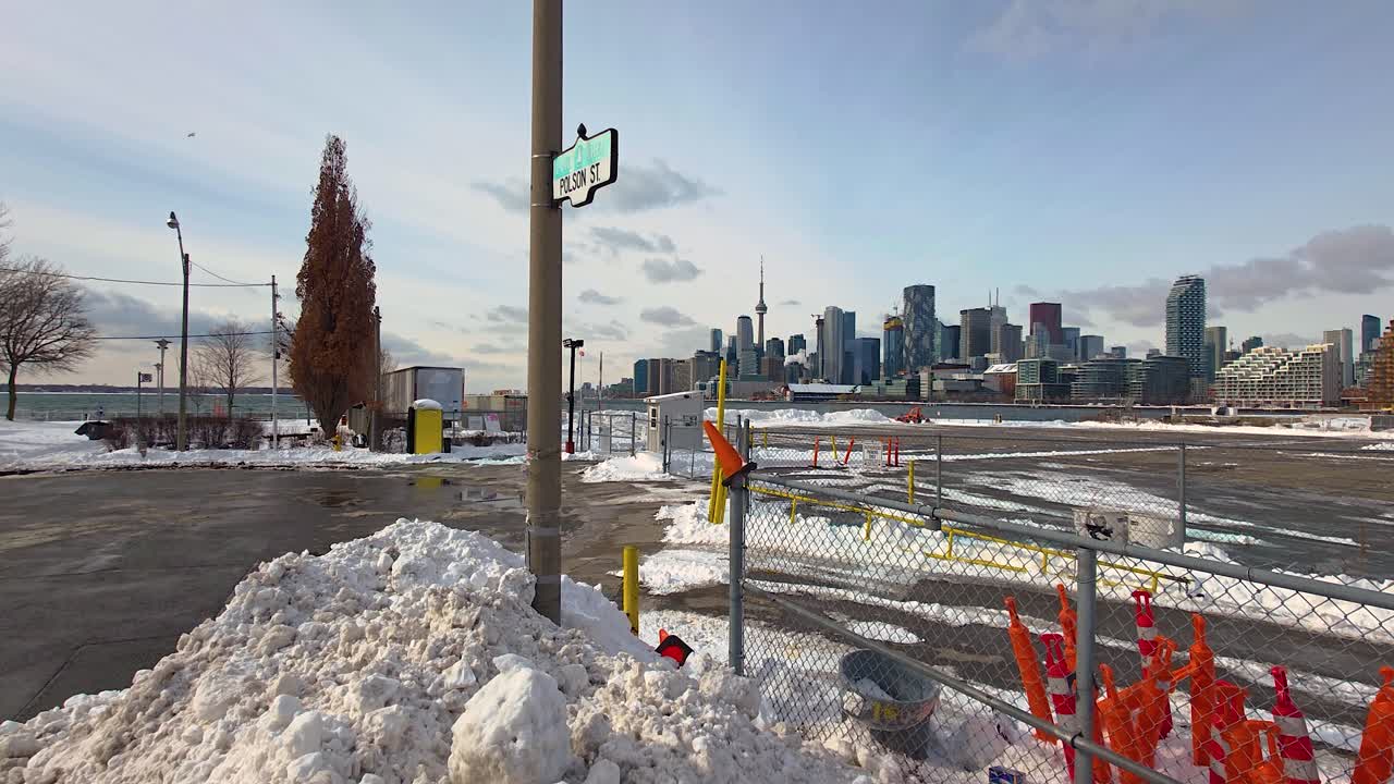 A scenic view of the Toronto skyline as seen from Polson Street, surrounded by snow banks and marked by orange construction cones, capturing the city's urban winter charm