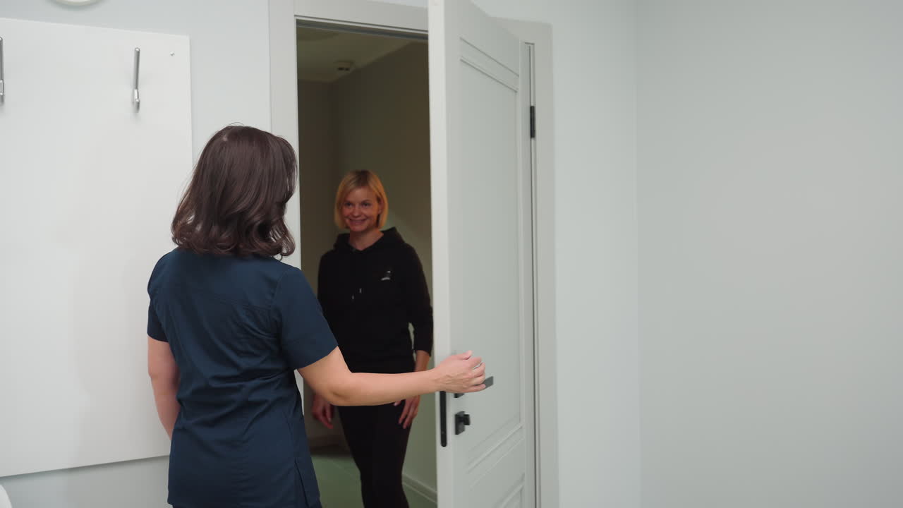 Cosmetologist in navy uniform opens door with warm smile while welcoming guest into bright, white painted room with minimalistic interior, and bright lighting, evoking professional hospitality
