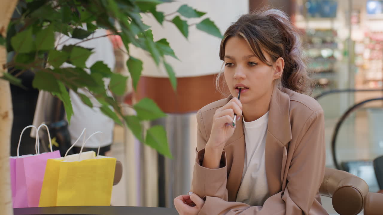 상점에서 립글로스를 평가하는 여성, 쇼핑 물품이 있는 소매점에서 립밤을 사용해 보는 젊은 여성, 쇼핑 액세서리를 주변에 두고 몰에서 베이지색 블레이저를 입고 립 제품을 검사하는 여성