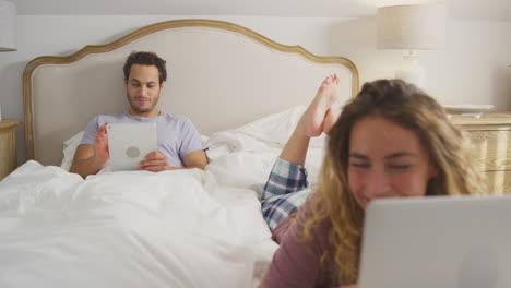 Pareja-En-Pijama-En-La-Cama-En-Casa-Con-Una-Mujer-Usando-Una-Computadora-Portátil-Y-Un-Hombre-Tableta-Digital