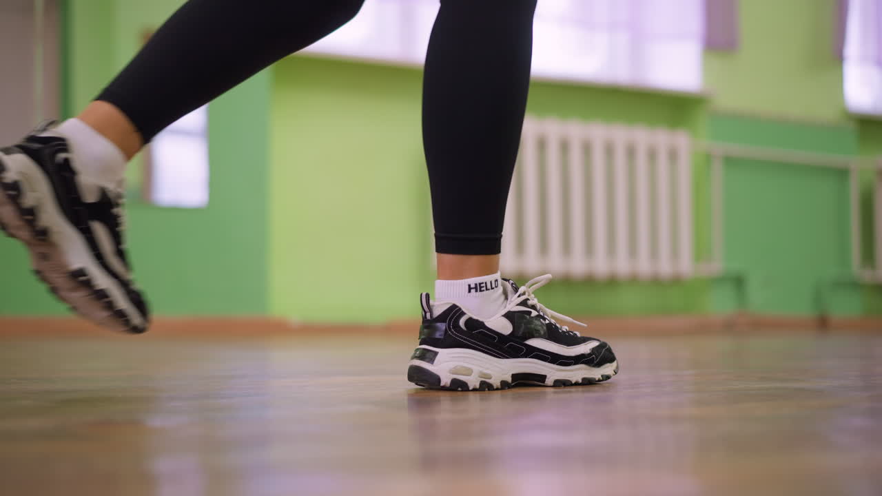 Leg view of woman in black leggings and canvas sneakers bouncing on polished wooden floor during indoor exercise, capturing rhythm, energy, and athletic motion with blurred green wall background