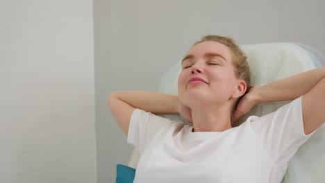 client with hair tied in bun reclines on chair, hands resting behind head while smiling softly. scene captures calm and relaxed posture with subtle expression in serene indoor setting