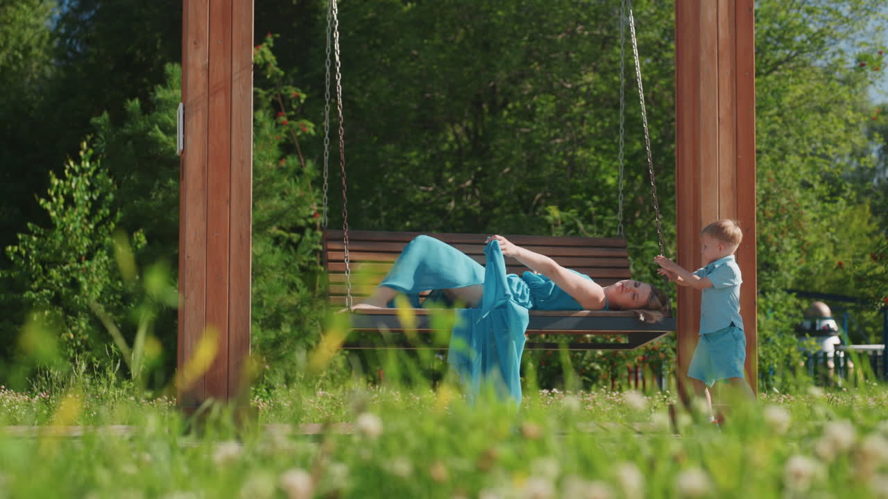Donna sdraiata su una panchina-altalena del parco mentre un bambino la tira e la stuzzica giocosamente durante una gita in famiglia in un soleggiato fine settimana. Un'interazione energica con erba, alberi, risate spontanee, una caduta improvvisa e gioiosa.