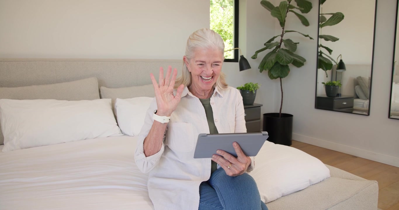 Senior woman sitting on bed at home, tapping tablet initiating call, smiling, waving, nodding