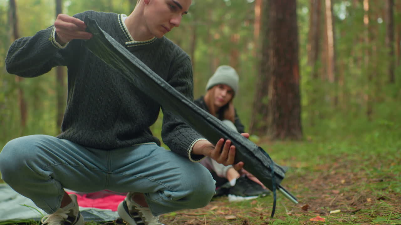 Young man squats in forest adjusting tent poles while female friend in gray beanie sits nearby watching him with admiration, both surrounded by trees and greenery