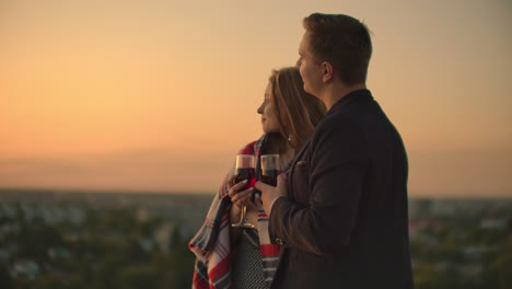 una joven pareja en el techo bebiendo vino de vasos de pie en una manta y admirando la hermosa puesta de sol sobre la ciudad.