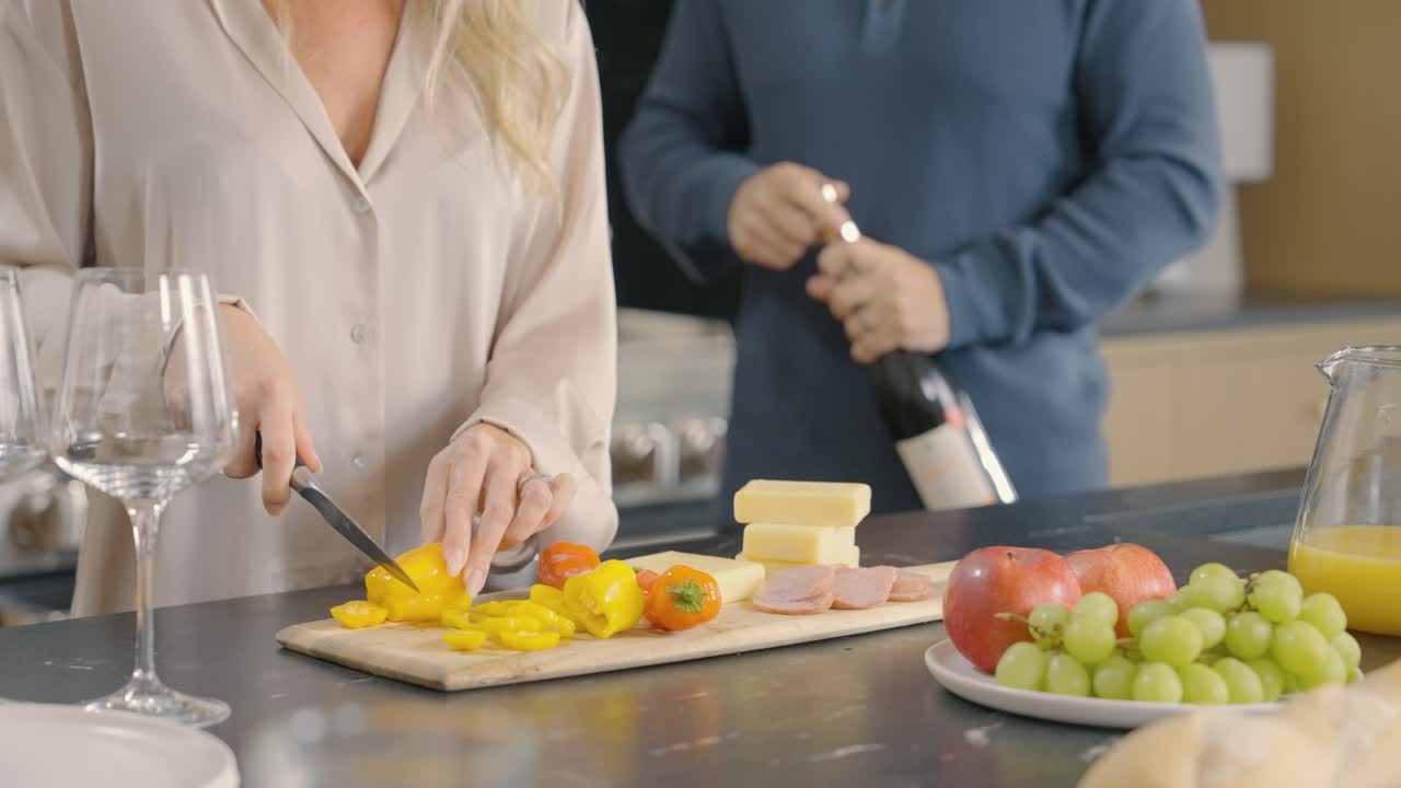 A woman is cutting vegetables, in preparing a charcuterie board while a man opens a bottle of wine in the kitchen