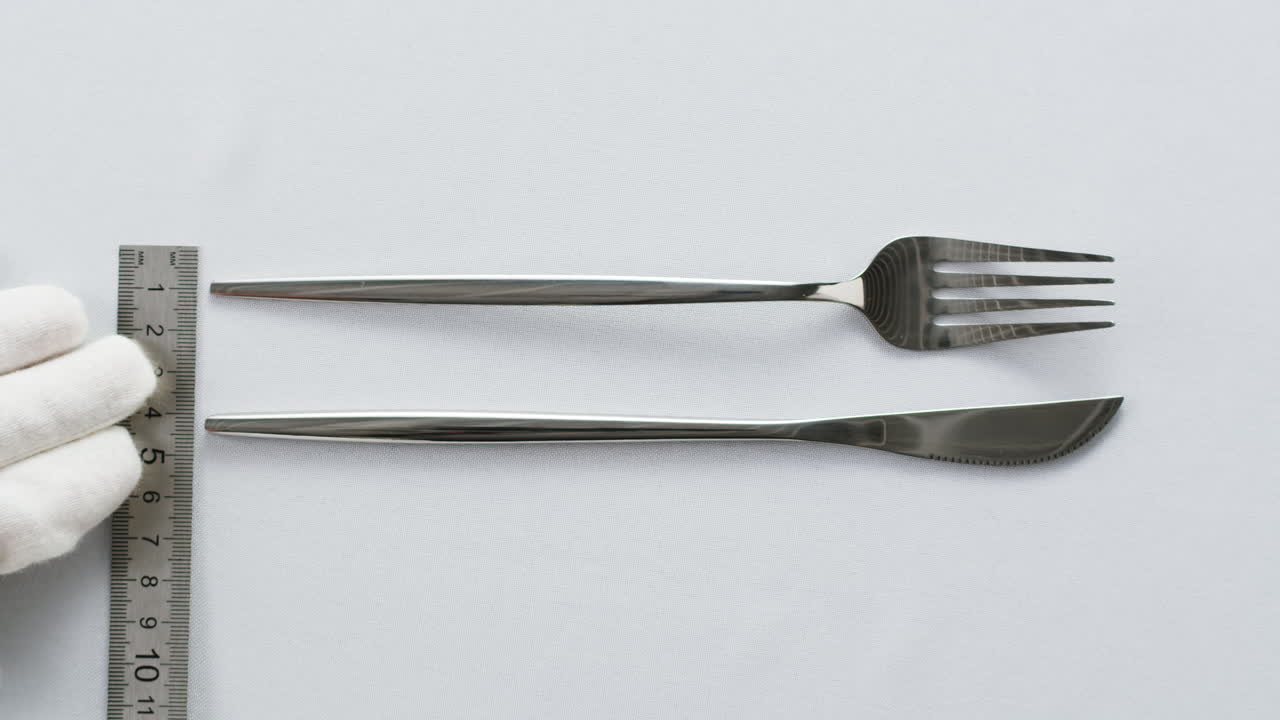 Minimalist table setting with white plate, fork, and knife arranged neatly, and a gloved hand adjusting cutlery.