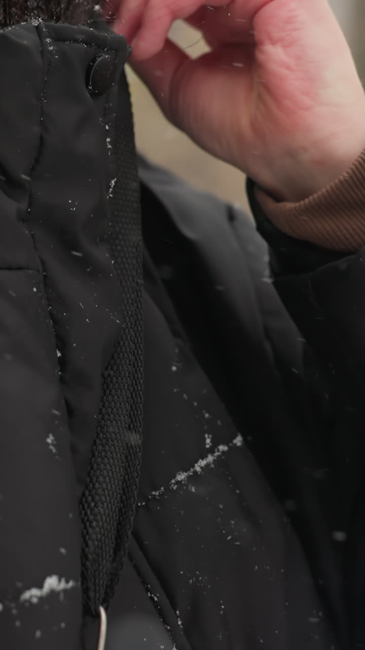 primer plano de una persona ajustando la correa de una chaqueta de invierno negra, copos de nieve visibles en la tela y el cabello, con árboles de otoño borrosos en un fondo de parque cubierto de nieve