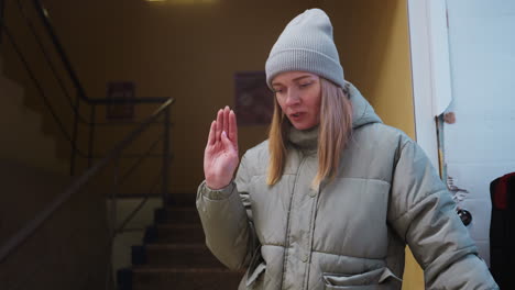 woman in green winter jacket and gray beanie stands indoors near staircase with serious expression, looking down while holding hand out, possibly commanding pet or engaging in quiet moment in old building