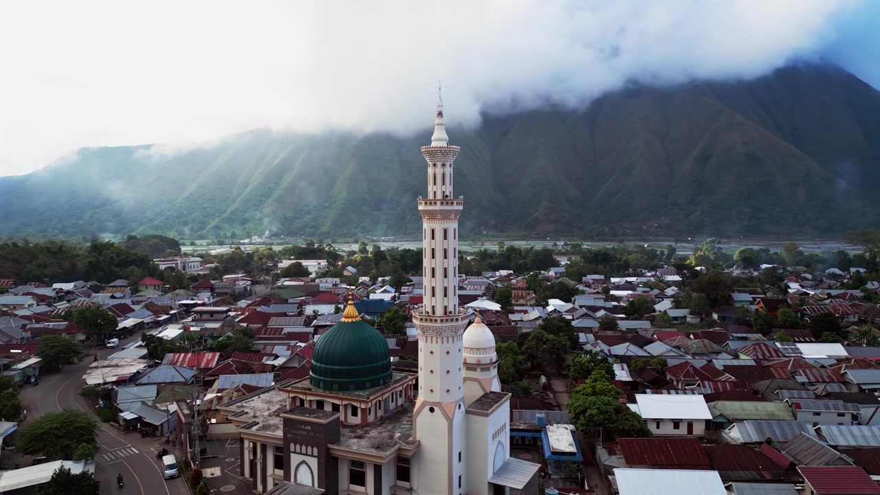 Desa pegunungan pedesaan berpusat di sekitar masjid agung dengan latar belakang pegunungan yang dramatis dan puncak yang tertutup awan di Indonesia, tampilan drone udara